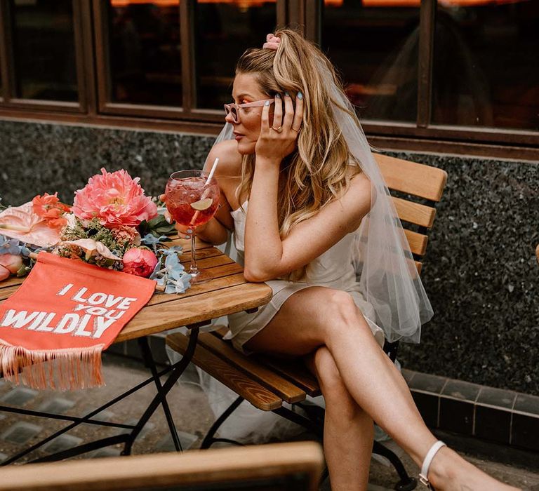 Bride in a retro pink sunglasses and short wedding dress drinking fruity cocktail with a fabric wedding banner on the table 