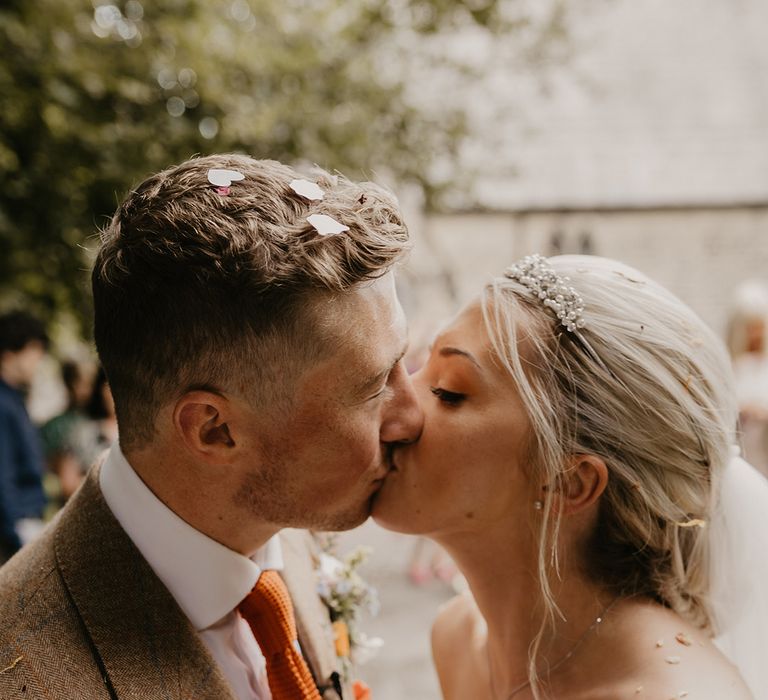 Bride in pearl tiara kisses the groom in brown suit and orange tie