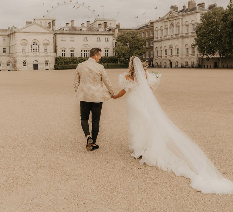 Bride in ruffle wedding dress with a long train walks with groom in custom tuxedo around their wedding venue in London 