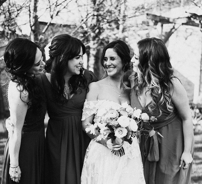 Bride in lace wedding gown smiles with bridesmaids in dark coloured dresses 