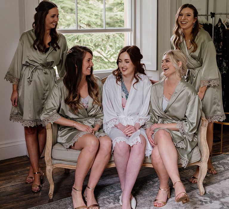 Bride in white satin robe getting ready with bridesmaids in green satin robes and matching gold sandals for wedding day 