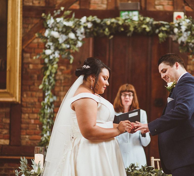 Bride with red wedding nails places the ring on the groom's finger who wears a three piece blue suit 