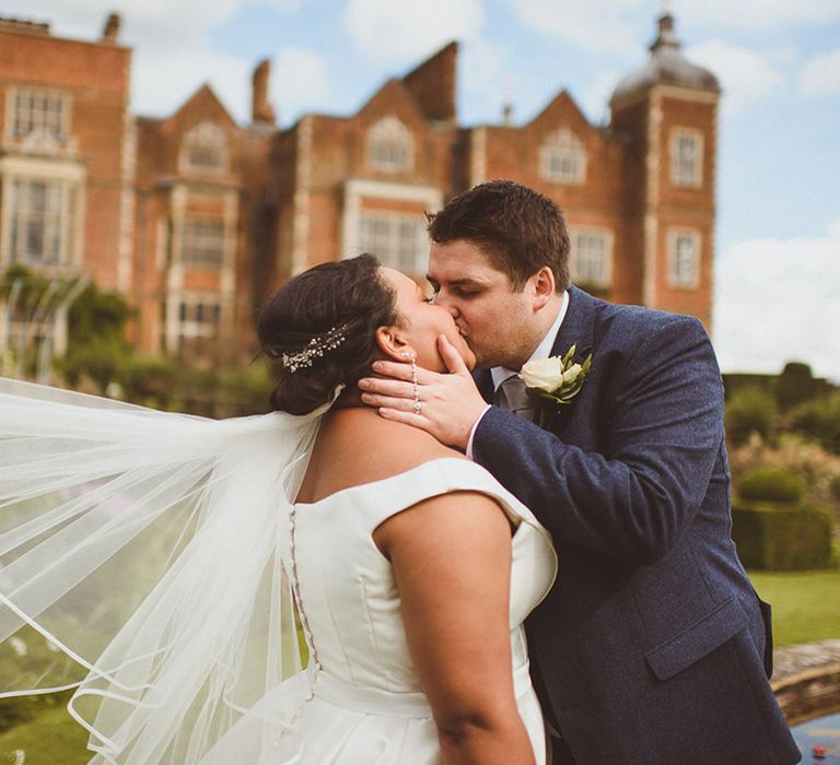 Bride and groom kiss in front of Hatfield House with bride's veil blowing in the wind 