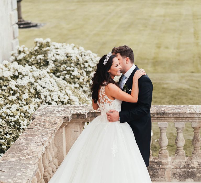 Bride and groom embrace on a balcony at Kirtlington Park House for royal inspired fairytale wedding 