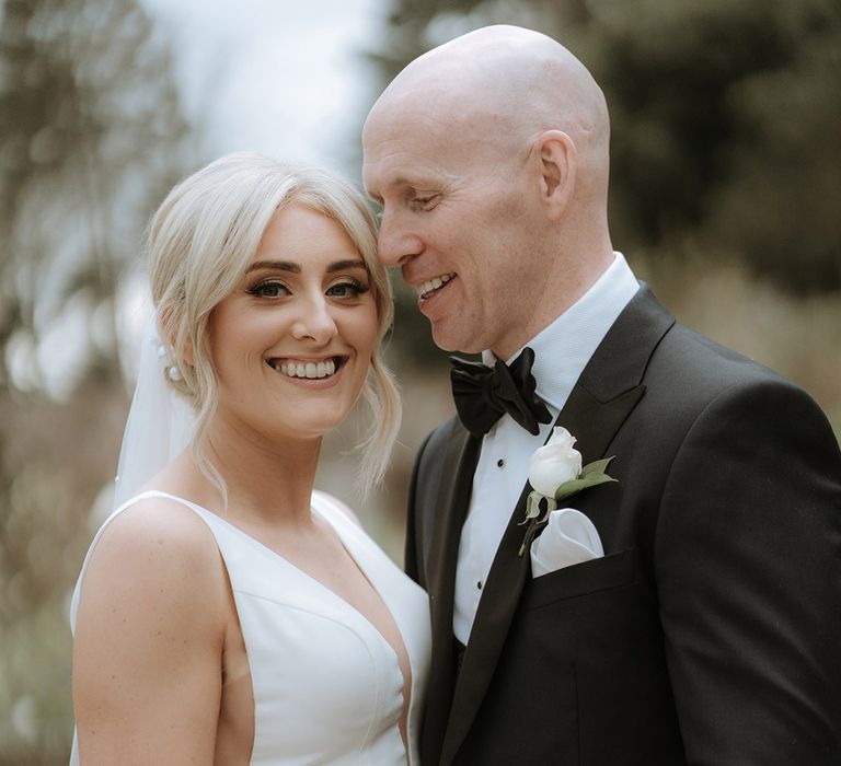 Groom in black tie smiles at the bride with bride in deep v fitted wedding dress 