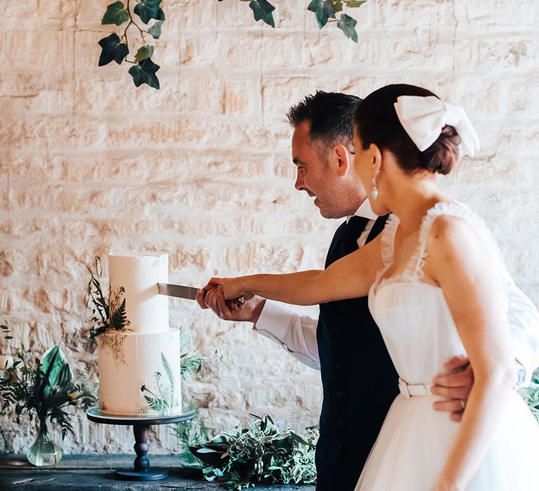 Bride with large white hair bow and ruffle tulle wedding dress cuts the cake with the groom in a blue suit 