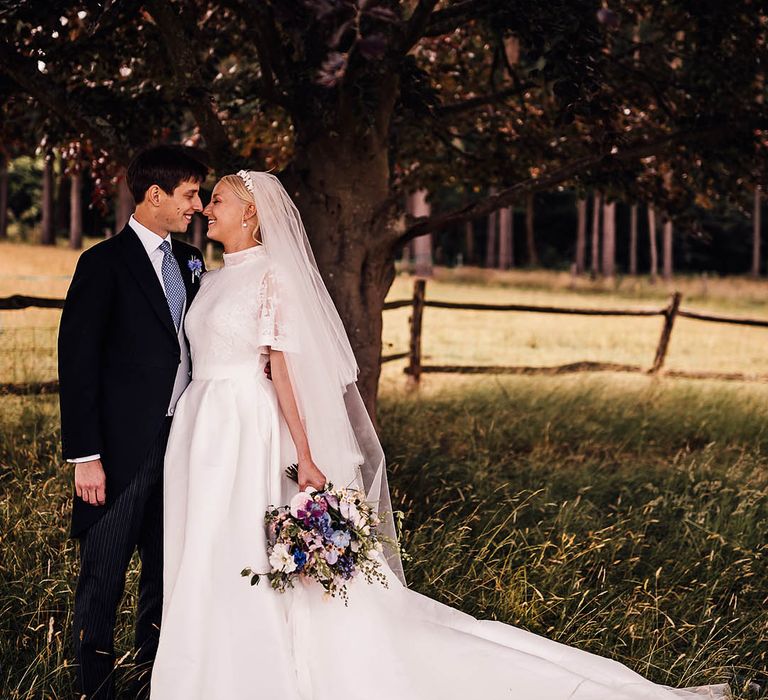 Groom in black morning sun with a blue tie and buttonhole stands closely with the bride in a lace wedding dress 