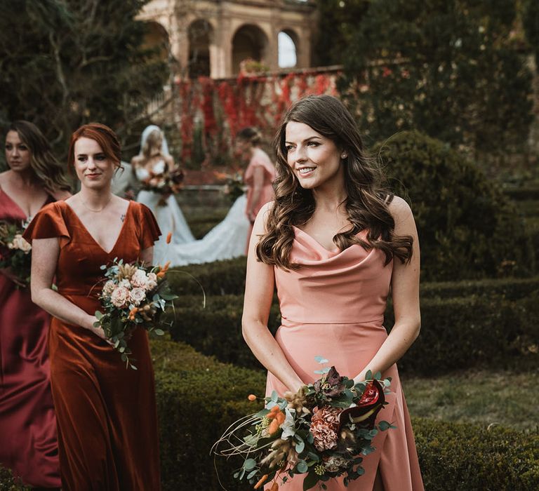 Bridesmaid walks down the aisle wearing different coloured orange dress complete with low cowl neck and thigh split to front 