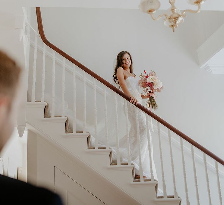 Bride in fitted lace wedding dress holding pink and white bridal bouquet walks down staircase and sees her groom for the first time on the morning of their wedding day