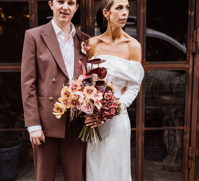 Groom in a brown suit with an unbuttoned shirt stands with the bride in a satin off the shoulder wedding dress holding a yellow, pink, and brown bouquet 