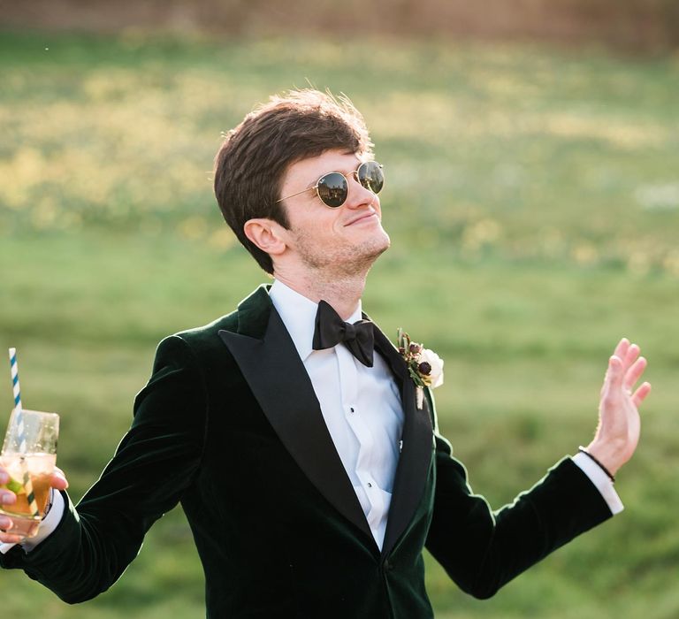 Groom wears black-tie and velvet green suit jacket with his sunglasses on his wedding day 