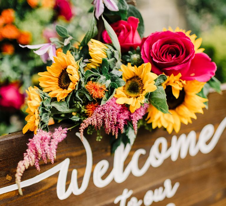 Wooden welcome sign with white writing surrounded by bright floral decor and statement sunflowers 