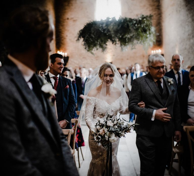 Father of the bride in a grey woollen suit walks the bride down he aisle at their rustic wedding 