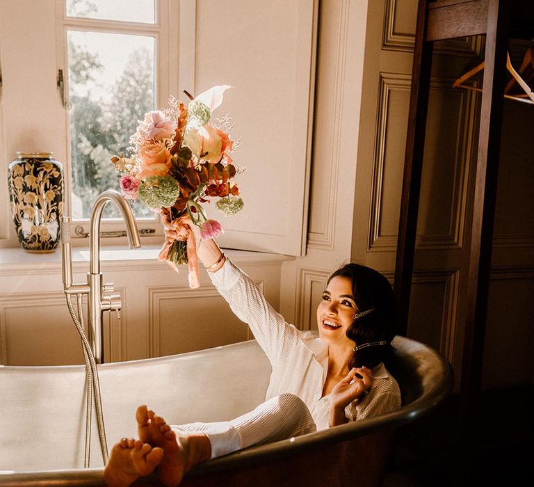 Bride in white bridal pyjamas in a copper bath tub holding her autumnal wedding bouquet 