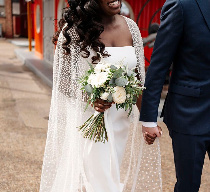 Black bride wears tulle wedding cape with glitter polka dots and holds classic white floral bouquet as she walks with her groom