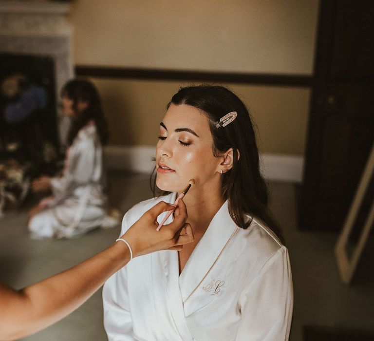 Bride sits in a white satin robe getting her makeup done for her big day 