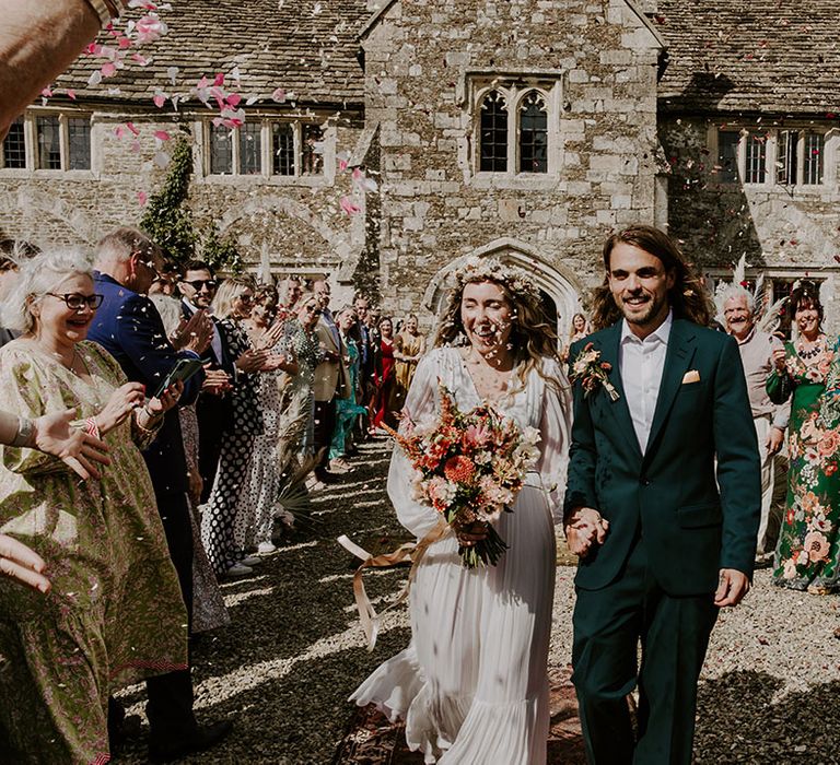 Groom wears green suit with floral buttonhole and pocket square and walks with bride carrying floral bouquet