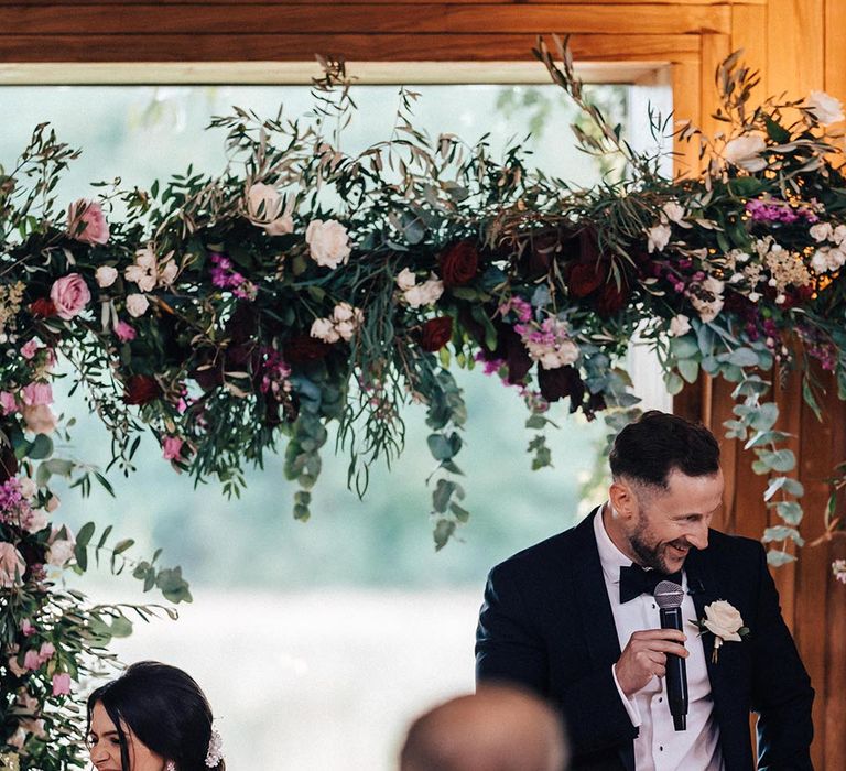 The groom in a black tuxedo stands with a microphone to read out his wedding speech as the guests smile and listen 