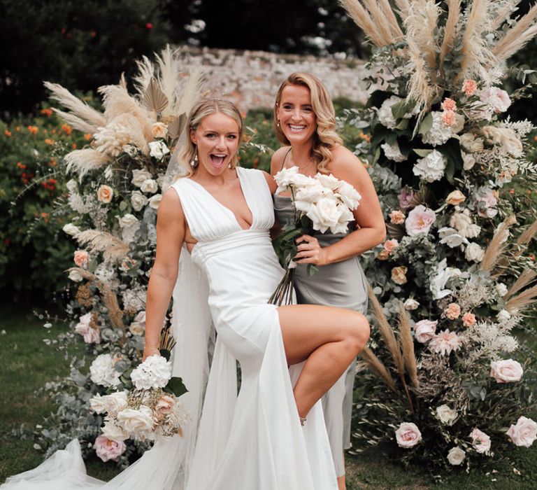 The bride and bridesmaid pose together in front of the flower columns with pink roses and pampas grass 