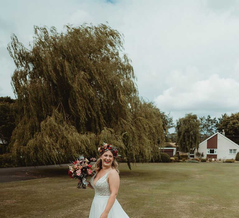 Bride wearing large flower crown holding colourful bouquet with bride in sparkly wedding dress with long white train 