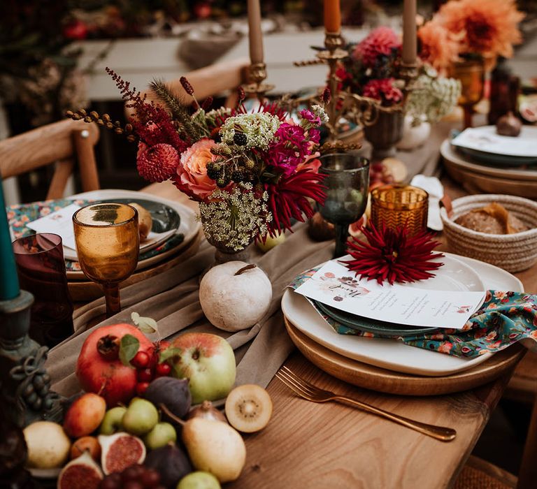 Neutral-toned tablerunner on rustic wooden table, orange, green and red coloured glasses, earthenware, seasonal fruit and vegetables, red, brown and green tapered candles in gold candlesticks and clear vases with autumnal coloured flowers