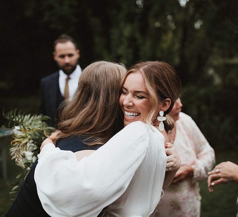 Bride wears statement earrings and long sleeve wedding dress and hugs wedding guest on the day of her wedding 