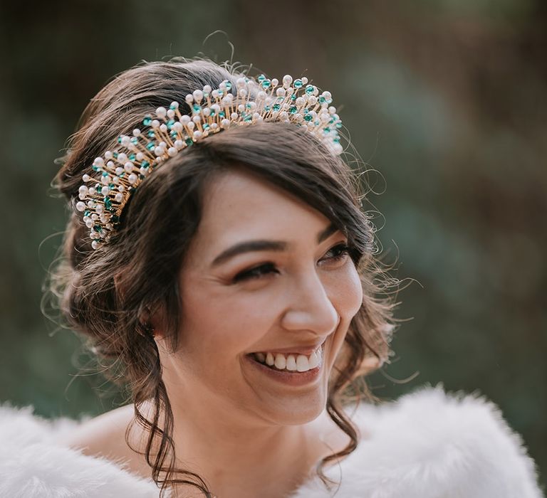 Bride in cropped faux fur bridal cover up and gold, pearl and emerald bridal crown smiling