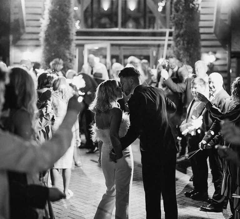 Bride in feather jumpsuit and groom in classic black tuxedo sharing a kiss at stylish wedding at Old Kent Barn