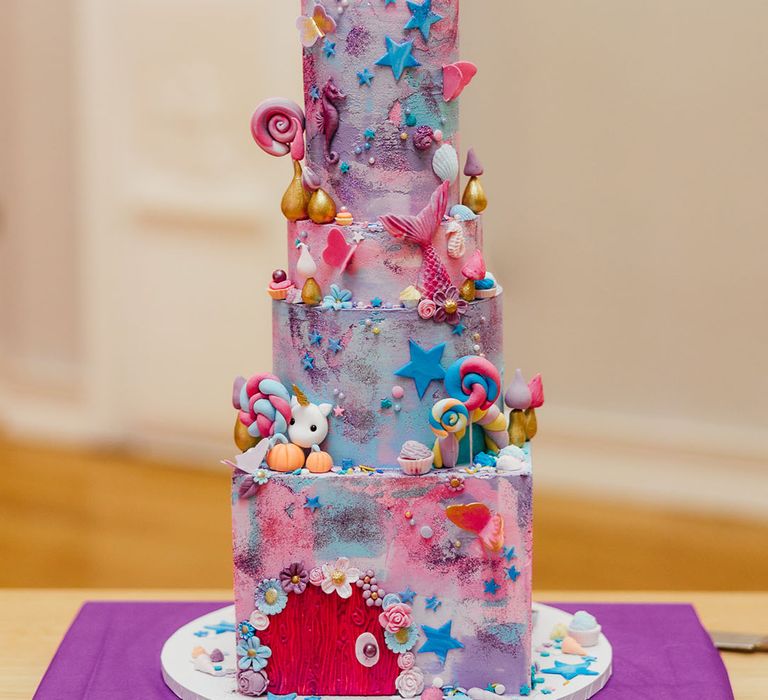 Four tier pink and purple iced wedding cake with cartoon and fairy decorations, lollipop, star, and flower icing and personalised pink and blue wedding cake toppers 