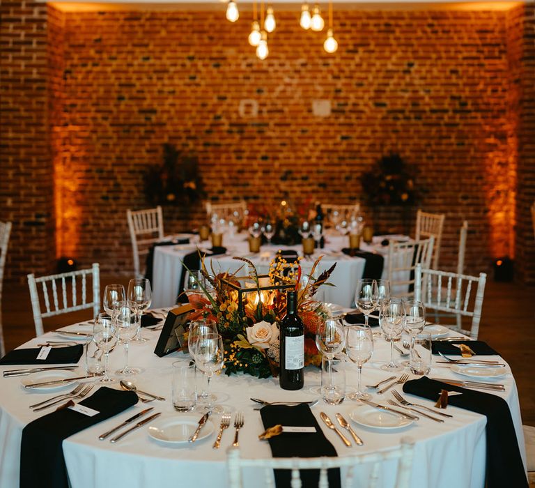 Round tables complete with white tablecloth and black fabric napkins alongside orange floral arrangements to the middle 