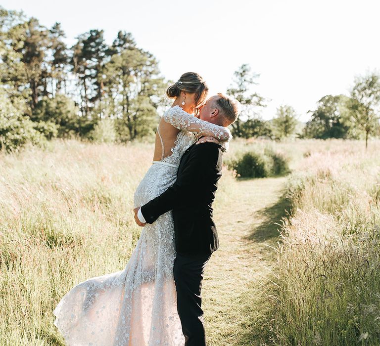 Groom in black tuxedo picks up and embraces the bride in a 3D lace sparkly long sleeve wedding dress with open back design during the sunset 