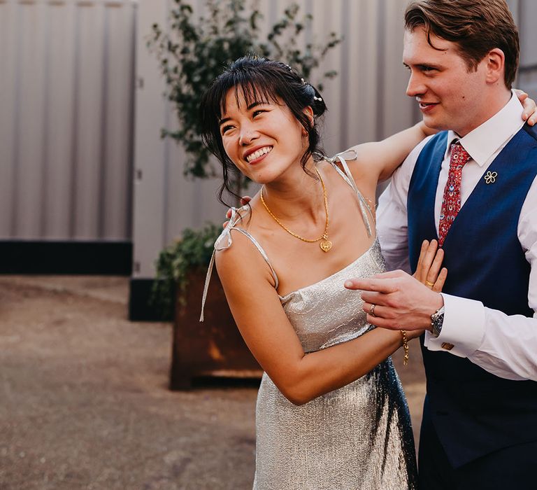 East Asian bride laughs beside her groom who wears white shirt and blue waistcoat complete with red tie 