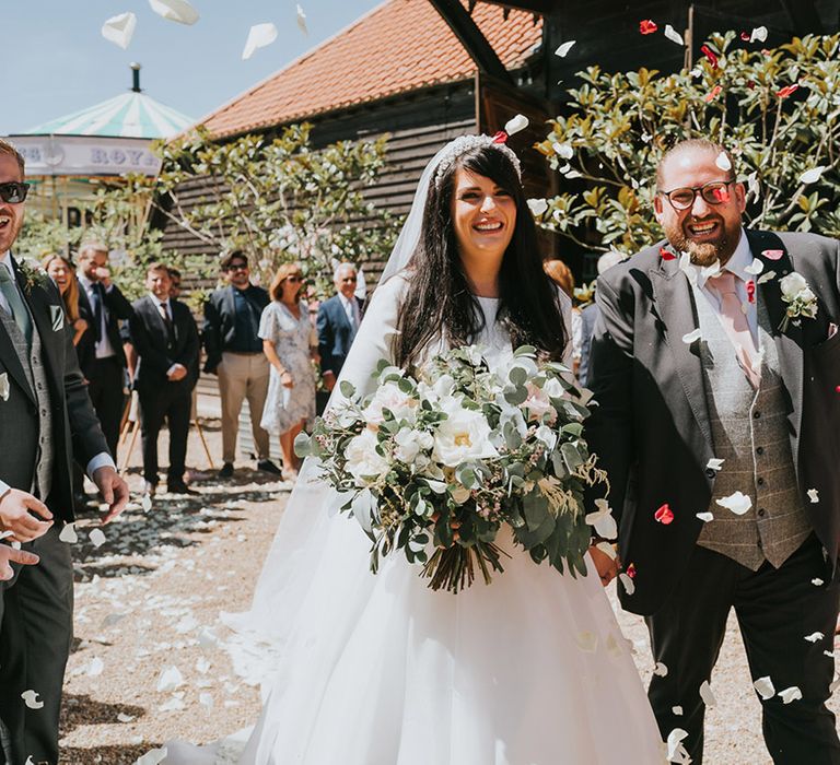 The bride wearing a tiara with a veil in a high neck long sleeve wedding dress has confetti moment with the groom in a dark grey suit jacket and light grey waistcoat 