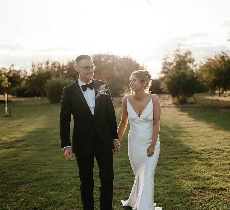 Groom in black tie and glasses walking during golden hour with the bride in a satin slip wedding dress 