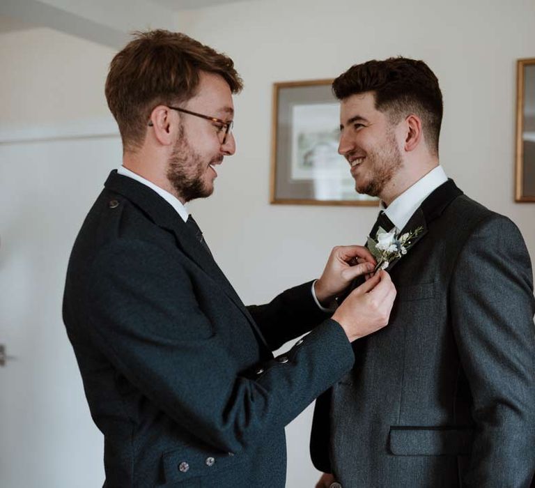 Groomsman helping groom in dark grey blazer and waistcoat and dark tartan tie with white flower boutonniere 