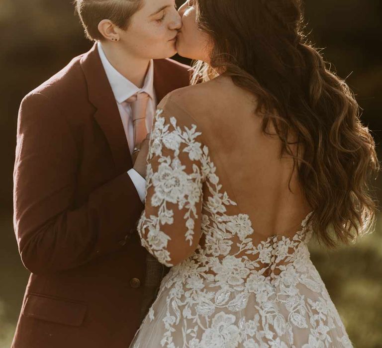 Bride in maroon bridal suit with grey waistcoat, dusky pink tie and autumnal dried flower boutonniere kissing bride in long sleeve lace illusion sleeve wedding dress with overlay at golden hour wedding photoshoot