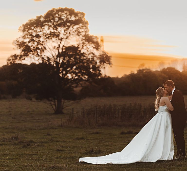 Bride in strapless Suzanne Neville wedding dress with long train posing with groom at golden hour 