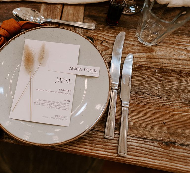 Wedding table place setting with white round plate, an orange napkin, silver cutlery and minimalist place name and menu stationery with bunny grass detail