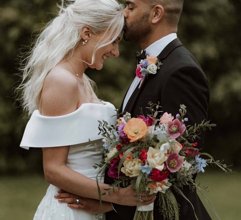 Bride in off the shoulder lace wedding dress holding colourful spring bridal bouquet standing with groom in classic three piece grooms tuxedo with colourful spring boutonniere at Frensham Hall wedding venue 