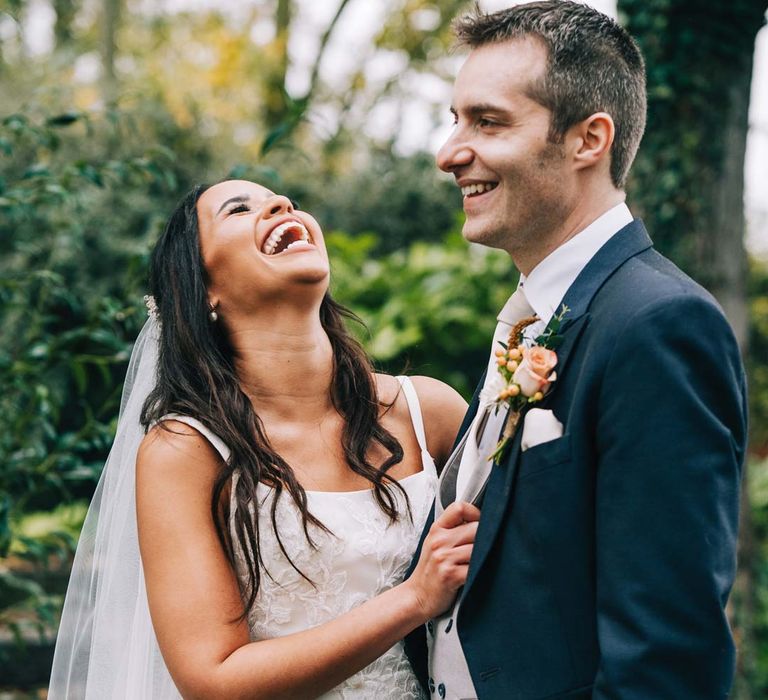 Bride in a-line tulle wedding dress with beaded lace applique with petals and a square neckline with side tulle cutouts laughing with groom in navy grooms morning suit with grey waistcoat, off-white tie and mixed wildflower boutonniere on grounds of South Farm wedding venue