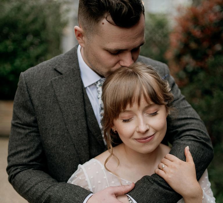 The groom in a grey suit embraces the bride in a dotted wedding dress from behind for special couple portrait 