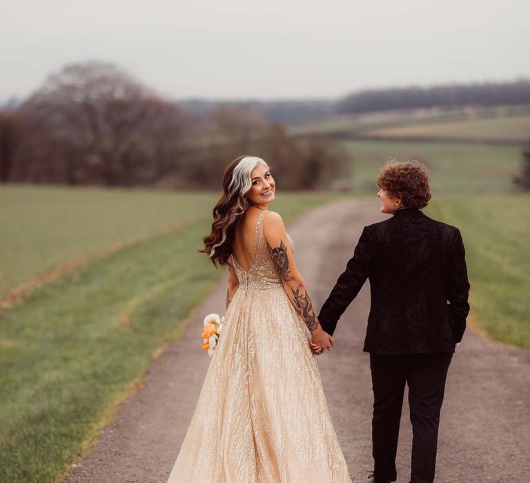 Bride looking back at camera in sleeveless deep v-neck sparkly gold wedding dress holding up orange garden rose, white poppy, light pink carnation and foliage bridal bouquet wrapped in sparkly gold ribbon with bride in dark bridal suit at Kingscote Barn Tetbury 