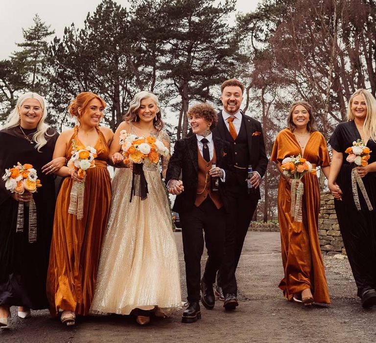 Bride in dark velvet bridal jacket with rust waistcoat and pocket square, black suit trousers, platform Dr Martens and gold pocket watch walking hand in hand with bride in sleeveless deep v-neck sparkly gold wedding dress with sheer and gold polka dot wedding veil holding orange garden rose, white poppy, light pink carnation and foliage bridal bouquet wrapped in sparkly gold ribbon with bridesmaids in black and orange mismatched bridesmaid dresses and groomsmen in dark three piece suit with orange tie and pocket square 