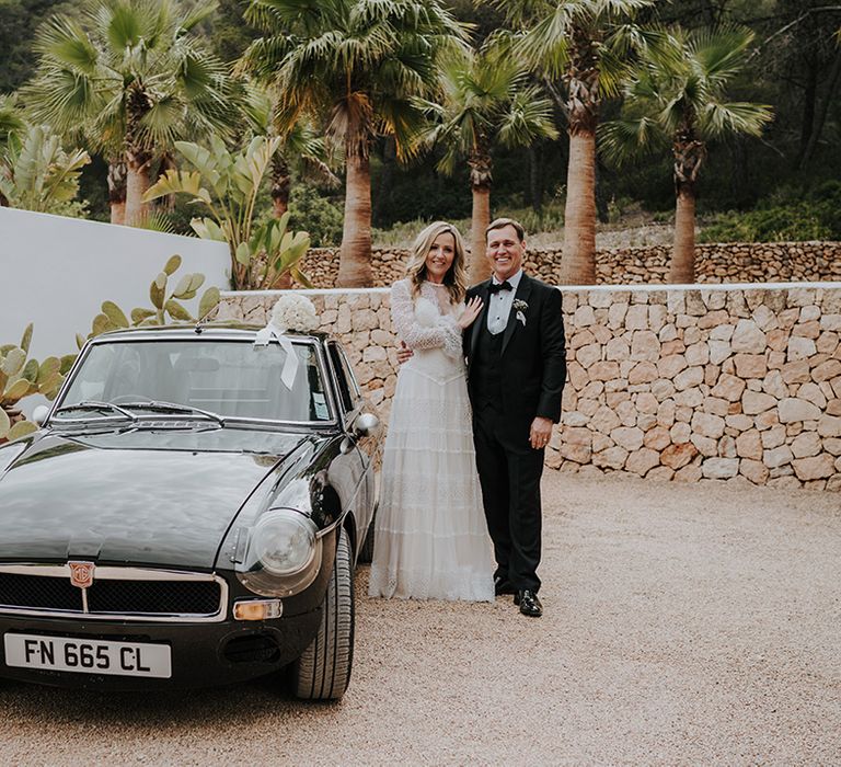 bride in a lace wedding dress with bell sleeves standing with her husband in a tuxedo next to a black vintage wedding car