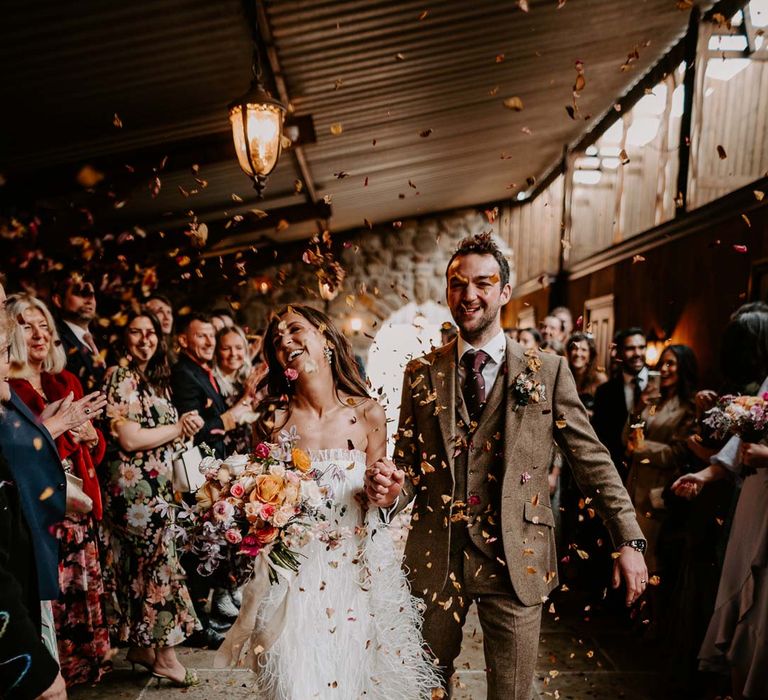 Bride in strapless ivory fringed crepe gown and chapel length veil holding mixed bridal bouquet with garden roses, carnations, peonies and foliage doing colourful dried petal wedding confetti exit with groom in chocolate brown grooms suit at Willow Marsh Farm Loughborough 