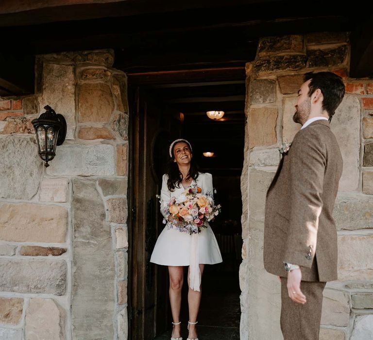 Bride in long sleeve short wedding dress, pearl bridal headband and platform pearl wedding heels holding mixed bridal bouquet with garden roses, carnations, peonies and foliage looking at groom in brown grooms suit 