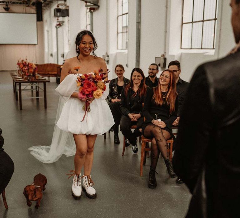 Bride in off shoulder tulle short wedding dress with church length veil and white platform Dr Martens wedding shoes holding vibrant pink and orange bridal bouquet walking down the aisle towards groom standing by vinyl wedding arch