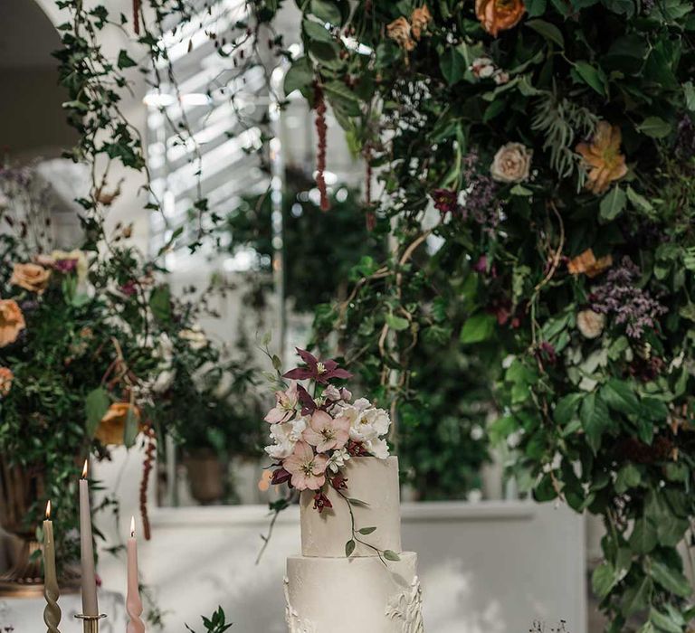 Four tier white wedding cake with intricate icing details and dried flower wedding cake topper on wooden table with twisted tapered candles in neutral shades in gold candlesticks surrounded by wildflower and foliage wedding flower arrangements