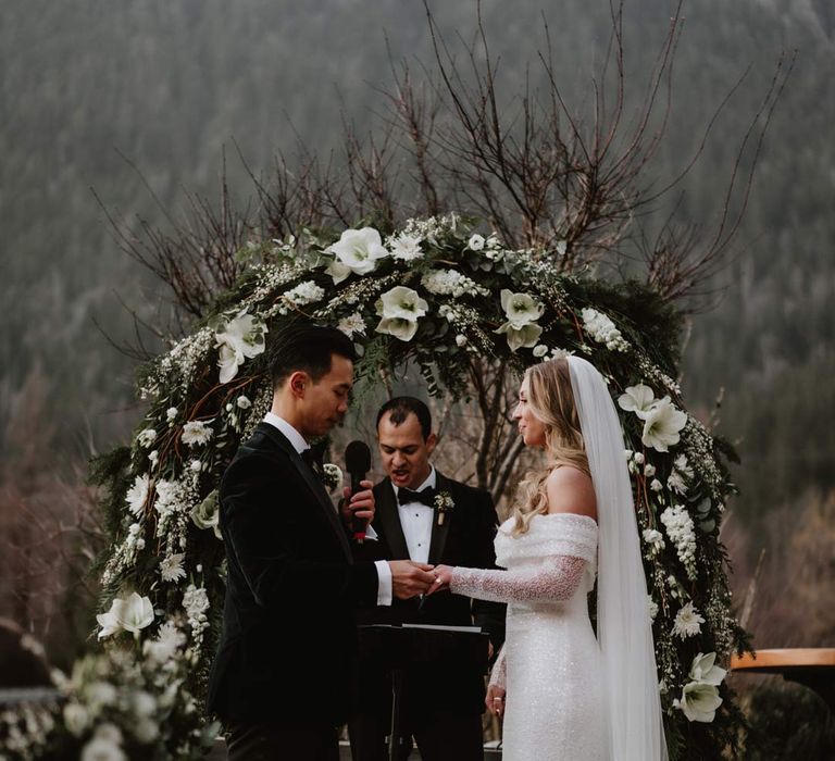 Groom in classic black tuxedo, black bowtie and white garden rose boutonniere holding hand of bride in off shoulder sparkly mermaid wedding dress in front of white garden rose, eucalyptus, baby's-breath and foliage wedding floral arch at French destination ski wedding