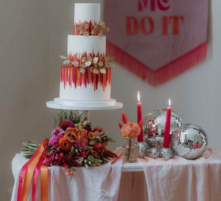 Three tiered iced wedding cake with pink and orange icing stripes and gold decorative leaves as cake toppers on table with dusky pink tablecloth, pink and orange steamers, disco ball wedding decor, pink tapered candles, and large retro pink and orange wedding banner 